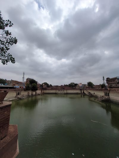 The image shows the Ghanta Ghar, also known as the Clock Tower, in Jodhpur, India. The sky is overcast with dark clouds. In the foreground, there is a large body of water, possibly a stepwell or reservoir, with brick structures lining its edges. The structures appear to be old and made of reddish-brown stone.
On the far side of the water, the Clock Tower complex is visible, featuring a tall tower with a clock face, surrounded by other buildings with traditional Indian architecture, including balconies and arched windows. Some of these buildings are painted in various colors, including blue and ochre. There are also some modern structures, such as a telecommunications tower, interspersed with the older buildings.
Along the edges of the water, there are steps leading down to the water level, and some people can be seen walking or sitting on the steps and along the embankment. There are also trees and vegetation growing in and around the structures. The overall impression is of an historic city center with a prominent landmark, captured on a cloudy day. The water is murky green, and there appear to be some debris floating on its surface.