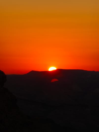 A serene sunset unfolds over the desert landscape of Yeruham, Israel. The sky is a vibrant gradient of warm oranges and reds, transitioning from a deep fiery hue near the horizon to a lighter, golden orange at the top. The sun, a glowing semi-circle, is positioned just above a dark, silhouetted mountain range that stretches across the middle of the frame. Below the mountains, the terrain fades into darkness, suggesting a deep valley or canyon. A dark, rocky formation juts out from the left edge of the image, further defining the foreground. The overall scene is one of natural beauty and tranquility, with the stark silhouettes of the desert contrasting against the dramatic colors of the sunset.