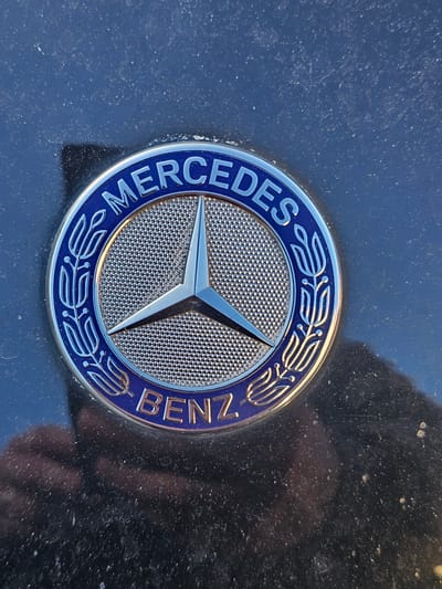 A close-up, slightly elevated perspective captures a Mercedes-Benz emblem mounted on a dark, glossy vehicle surface, likely a car hood or trunk. The circular emblem features a prominent silver, three-pointed star at its center, set against a textured, dimpled silver background. The star itself is reflective, showing highlights. Surrounding the central star, the outer ring of the emblem is dark blue, emblazoned with the silver text "MERCEDES" on the left and "BENZ" on the right. Stylized silver laurel wreath patterns adorn the blue ring between and around the text. The emblem appears clean and polished, contrasting sharply with the surrounding dark surface of the car, which is noticeably covered in a layer of fine dust and small particles. Reflections are visible on the car's surface, with a faint, distorted human-like shape, possibly the photographer's reflection, noticeable in the bottom right corner of the image. The lighting indicates an outdoor setting, possibly daylight, illuminating the emblem's metallic sheen. The scene is located in Holywell, United Kingdom.