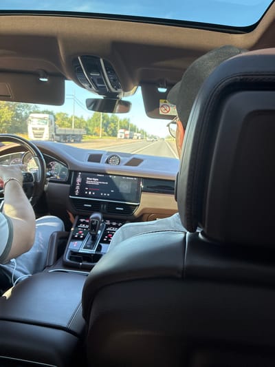 The image captures the interior of a modern car, likely a Porsche, as seen from the perspective of a rear passenger. The driver, wearing glasses and a black cap, is operating the vehicle on a sunny day. The car's dashboard displays a digital speedometer and a touchscreen infotainment system, currently showing music details for "Silver Night." A sun-visor is lowered on the driver's side, revealing an airbag warning sticker. Outside the car, visible through the windshield and sunroof, is a rural or suburban landscape with trees and a road lined with power lines. Several trucks, including a white Volvo, are driving on the road. The scene is consistent with a drive through the outskirts of Podol'e, Russia, where such road conditions and vehicle types are common. The interior is well-lit, highlighting the car's luxurious finishes, such as leather seats and a panoramic sunroof.