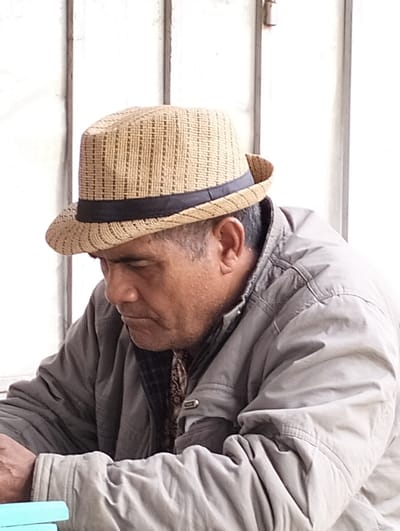 A man wearing a light brown woven hat with a dark ribbon sits in front of a white, vertically paneled wall. He wears a gray jacket, partially covering a patterned shirt underneath. His head is tilted down, and his gaze is directed towards the bottom left of the frame. The man's hands rest on a stack of blue objects. The lighting is soft, with subtle shadows defining the folds of the jacket.