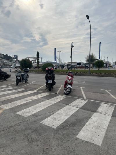The image displays an outdoor asphalt street scene in L'estartit, Spain, under an overcast sky. In the foreground, a worn white pedestrian crosswalk is visible on the asphalt. Three scooters are parked in the midground: a light grey scooter facing left, a black scooter with a red top case facing away from the viewer with license plate B 822 LCK visible, and a red and white scooter facing right. Part of a dark motorcycle is also visible on the far left. A single cigarette butt lies on the asphalt near the crosswalk. In the background, beyond a landscaped median with bushes and small rocks, a marina is discernible. The marina includes numerous boats, a large white gantry crane, and several associated buildings. Two tall blue banners, partially obscured, bear white text including "SAILING SCHOOL." Hillsides dotted with white buildings are visible on the far left. No individuals are present in the scene.