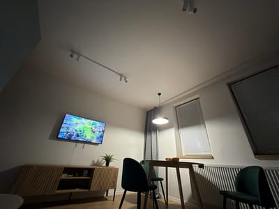 The image depicts an indoor scene, likely a living room or dining area, with a noticeable slant to the floor or walls. On the left, a modern, slatted wooden cabinet sits beneath a flat-screen television mounted on the wall, displaying what appears to be a video game. To the right, a wooden dining table with two dark green chairs is visible. A sheer grey curtain hangs behind the table, and a window with blinds is situated to the right of the curtain. The ceiling features track lighting and a pendant lamp. There is a radiator visible along the bottom right of the frame. The overall lighting suggests it is either nighttime or the room is dimly lit. There is no indication of the city of Sosnowiec, Poland, within the image itself.