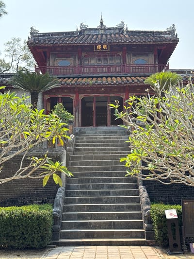 The image shows a multi-tiered Asian building with a traditional roof and intricate ornamentation, situated at 99 Kim Long, Kim Long Ward, Thua Thien Hue, Vietnam. The building has red accents and features decorative elements on its façade, including Chinese characters on a plaque. In the foreground, a concrete staircase leads up to the building's entrance, flanked by greenery and small bushes. A slender tree with green leaves partially obscures the view of the building and staircase. The environment suggests a historic or cultural site.