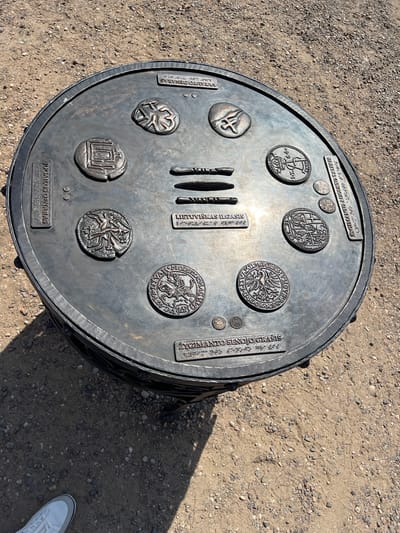 The image shows a circular, bronze monument in the city of Trakai, Lithuania, placed on a gravelly ground. The monument depicts various historical coins and symbols, possibly representing the monetary history of Lithuania. Inscriptions in Lithuanian and braille are visible on the monument's surface. The monument appears to be a public art installation or a historical marker. The background consists of dry, sandy soil with scattered pebbles and some shadows cast by the monument. No people are clearly visible in the image.