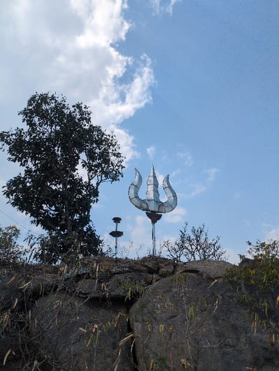 The image depicts a static scene on a rocky elevation in Kadaladi, India, under a blue sky with scattered white cumulus clouds. A prominent, large, metallic silver-colored trident (Trishula) is vertically mounted on a pole, situated near the center-right of the frame, rising above a rock formation. To its left and slightly behind, a smaller, dark metallic structure, cylindrical with two horizontal tiers, is also mounted on a pole. A large tree with dense, dark green foliage occupies the left side of the frame, partially silhouetted against the sky. The foreground and midground are dominated by substantial grey rock formations, exhibiting rough textures and visible cracks, with sparse, dry, light-colored herbaceous vegetation growing amongst them. No individuals or animals are present. No actions or interactions are observed.