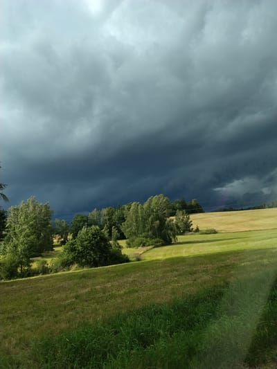 The image depicts a rural landscape in Ježená, Czechia, dominated by a dramatic sky. Dark, heavy storm clouds fill the upper portion of the frame, suggesting an impending storm. Below the clouds, a line of lush green trees runs horizontally across the midground. Beyond the trees, a field of golden, recently harvested crops stretches into the distance, contrasting with the vibrant green of the grassy foreground. The overall impression is one of nature's power and the impending arrival of stormy weather. There are no people or specific man-made structures visible in the scene.