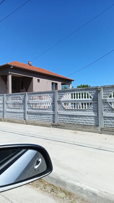 The image shows a residential scene in Boliartsi, Bulgaria. A light brown, two-story house with a red tile roof is partially visible behind a long, ornate gray concrete fence. The fence has decorative openings with vertical bars and oval shapes. Overhead electrical wires are strung against a clear blue sky. A side view mirror of a car is in the lower left corner, suggesting the picture was taken from a moving vehicle. The street in front of the fence is paved and clean. No people are visible. The scene is brightly lit, indicating a sunny day. Vegetation is sparsely visible in the background beyond the house.