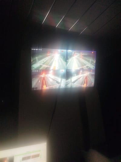 The image shows a dark room with a large monitor displaying a four-way split screen of CCTV footage from a highway at night. The footage shows traffic lanes with illuminated barriers and overhead lights. There are digital timestamps on each feed. The location is identified as Түндүк айланып өтүүчү жол 84 га, Kyrgyzstan. A portion of a white computer monitor or control panel is visible in the lower left corner, displaying some data and controls, with a bright light emanating from it. The overall environment appears to be a control room or monitoring station.