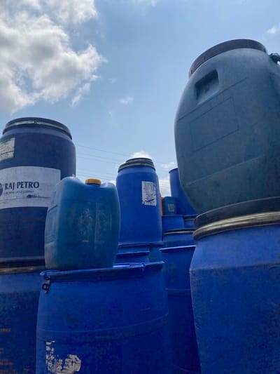 The image shows a stack of blue plastic barrels under a partly cloudy sky. The barrels are of varying sizes and some have visible labels. One barrel in the foreground has a white label with blue text that reads "RAJ PETRO" and below it, "A BRENTAC GROUP COMPANY". Another barrel has a yellow cap. The barrels appear to be outdoors, possibly in a storage area or an industrial setting in Amaseri, Nigeria. The lighting suggests it is daytime, and the sky indicates fair weather with scattered clouds. The perspective is from below, looking up at the stacked barrels and the sky. There are no people visible in the image.