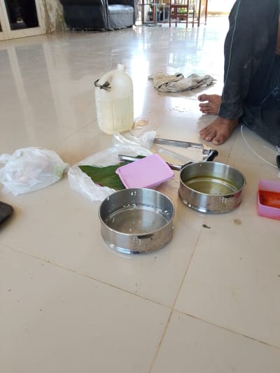The image depicts a tiled floor indoors, possibly a covered patio or room with an open side, given the natural light. The scene includes several objects laid out on the floor: a large, translucent plastic jug with a handle containing a pale liquid, a crumpled white cloth, a pair of metal tongs, two round stainless steel containers (one with some white residue, possibly rice, and the other with a yellowish liquid), a pink plastic container with a lid, and a pink rectangular plastic container holding an orange substance. To the left, there are two white plastic bags, one containing some items and the other appearing empty. A person's bare feet, with visible toenails, are visible in the lower right portion of the image, suggesting they are seated on the floor. The overall environment appears somewhat unkempt, with water or liquid spills on the floor and some scattered debris. The context suggests a casual or informal setting, possibly related to food preparation or a meal.