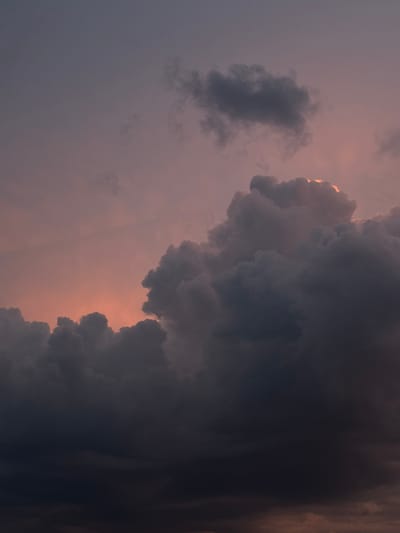 A vertical, close-up photograph captures a dramatic sky filled with dark, billowing clouds during twilight. The lower portion of the image is dominated by deep charcoal and violet hues, suggesting thick cloud cover. As the eye moves upward, the clouds become lighter, revealing streaks of dusky pink and orange from the setting sun, visible between the cloud formations. A few lighter, wispy clouds are scattered in the upper background, transitioning into a muted purple sky. The scene is devoid of any human presence, animals, or man-made structures, focusing solely on the atmospheric conditions. The location is identified as Savenki, Russia, implying this is a natural landscape vista from that region.