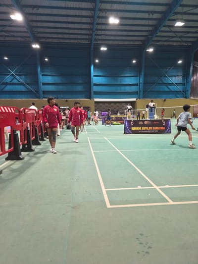 The image depicts the interior of a gymnasium, likely hosting a sporting event, possibly badminton or volleyball, as indicated by the court markings. In the foreground, two young individuals wearing matching maroon athletic outfits and knee pads are walking across a light green court. To the left, a red barrier with white stripes separates the court from a walkway. In the background, several other individuals, some in athletic wear, are visible, including a person seated on a high umpire's chair. A large banner with text "KENDRIYA VIDYALAYA SANGATHAN 55th NATIONAL SPORTS MEET 2026" is displayed on a dark background. The gymnasium has a high, corrugated metal roof supported by blue beams, and illuminated by overhead lighting fixtures. The overall environment suggests an organized sports competition within an indoor facility. The scene is located in Phagwara, India.