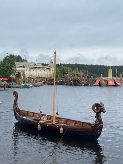 In the city of Моторное, Russia, on a cloudy day, a Viking longship with a carved ram's head prow is moored in a body of water. The ship is dark wood with intricate carvings along its hull and is equipped with a single mast and rigging. In the background, a large, fortified wooden structure resembling a castle or settlement sits on the shore, with several smaller boats and platforms visible around it. The sky is overcast, and the water is calm, reflecting the surrounding structures and the ship. The scene suggests a historical reenactment or a themed attraction.
