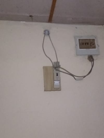 The image depicts an electrical setup on a wall in Pantinapu, Nigeria. The primary subjects are two electrical outlets and wiring. One outlet is a single socket with a brown interior, mounted on a white rectangular plate attached to the wall. This outlet appears to be an older model. A bundle of grey wires originates from this outlet, connecting to a junction or connector block.

Below and to the right of the first outlet is a second electrical unit. This unit features a switch and possibly another type of connector, mounted on a light brown wooden base that is itself attached to the wall. A grey wire runs from the first outlet to this second unit, and another grey wire emerges from the second unit and disappears off-camera. A small, circular grey conduit is visible on the wall to the left, from which another grey wire emerges and connects to the first outlet.

The wall is a neutral, light color, likely painted. The overall impression is one of utilitarian, potentially older or temporarily installed electrical infrastructure. There are no people visible in the image. The lighting suggests an indoor setting, possibly during the day. The scene appears to be a close-up view of the wall, focusing on the electrical components. No discernible text is visible in the image. The context provided indicates this is located in Pantinapu, Nigeria, suggesting a residential or commercial building in that region.