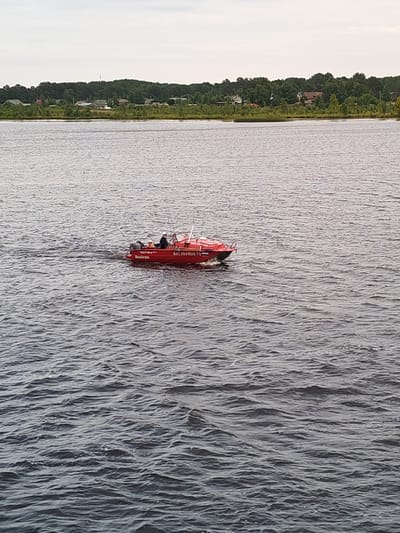 A red boat with three people on board is moving across a body of water, likely a river, near the city of Shal'skii, Russia. The boat has text written on its side, including "AH2 494 RUS 78". The water is choppy, and in the background, a tree-lined shore with scattered houses can be seen under a cloudy sky.