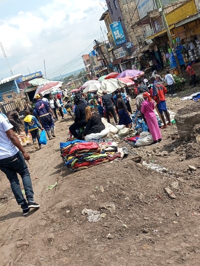 This image depicts an active, unpaved market street in Keekonyokie ward, Kenya, under a partially cloudy sky. The ground consists of uneven dirt with scattered debris and small mounds of earth. Multi-story buildings, appearing utilitarian and displaying commercial signage such as "MOZZART BET" and a blue billboard for "Gold Crown" milk, line the background. Numerous individuals, identified as vendors and shoppers, populate the scene, standing, walking, and interacting within the market space. Large, colorful market umbrellas provide shade over various informal stalls where goods are displayed. In the mid-ground, a prominent pile of folded, colorful clothing is laid out on a dirt mound. A person wearing a white t-shirt and blue jeans walks in the immediate foreground, with their back to the camera. Another individual, dressed in blue overalls and black boots, walks away from the viewer, carrying a blue bag. A person in a bright pink dress or robe stands near a clothing pile in the mid-ground right. The overall environment suggests a bustling, open-air commercial area with informal setups.