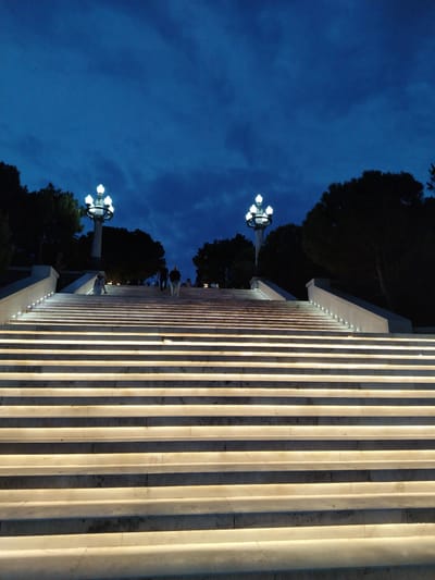 The image shows a wide, illuminated stone staircase in Sabail Raion, Azerbaijan, at twilight. The sky is a deep blue with some cloud cover. Two ornate streetlights with multiple bulbs are visible on either side of the upper part of the staircase, casting a warm glow. Several people are ascending the stairs; two figures are walking together further up, and one individual is visible on the lower half of the stairs, facing away from the camera. The steps are made of light-colored stone and are illuminated by embedded lights along their edges. Trees form a dark silhouette against the sky on the left and right sides of the scene. The architecture suggests an outdoor public space, possibly a park or monument entrance.