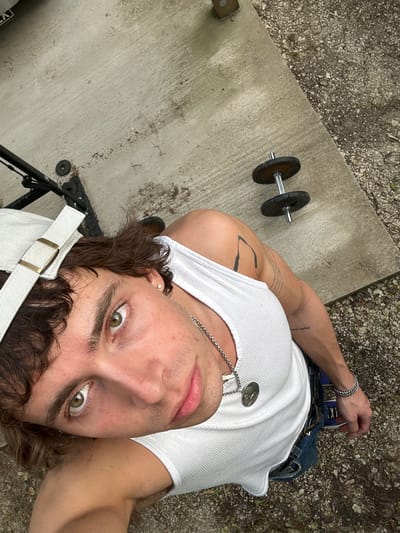A medium-angle, high-angle selfie shows a young male in a white tank top and blue jeans, lying on his back on a concrete surface outdoors. His head is tilted back. He wears a silver chain necklace with a coin pendant and a silver bracelet on his left wrist. He has dark curly hair and light eyes. A white hat with a gold buckle and strap is partially visible at the bottom of the frame.

To the left of the frame, a dumbbell lies on the concrete. The ground adjacent to the concrete is covered in gravel. A black metal structure is at the bottom left.

The location is unknown. The city of Lourmarin, France, is not discernible from the image.