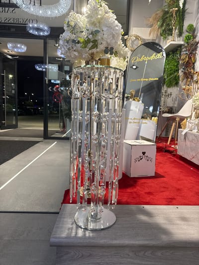 The image shows a store window display in Ilidža, Bosnia and Herzegovina. The display features a tall, crystal-covered stand adorned with white flowers, and a black, arched mirror with text in gold. Behind the display, the interior of the store is visible, with shelves filled with decorative items, including plants, gold accents, and a white chair. A red carpet covers the floor in front of the display. The entrance to the store is also visible, with glass doors and reflections of chandeliers and lights. The scene appears to be set up for a special event, possibly a celebration or a wedding.