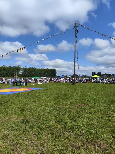 A bright, sunny day in NovoUrsaevo, Russia, is depicted in this image, with a festival or outdoor event taking place on a large grassy field. A tall wooden pole in the center of the frame supports a string of colorful triangular flags that stretch across the sky. The flags are attached to the pole at various heights, creating a festive and dynamic visual element.

A crowd of people is gathered, seated and standing, watching an event that appears to be a wrestling match or a similar athletic competition. A wrestling mat, a large rectangle with yellow and red markings, is laid out on the grass in the foreground. Spectators are arranged in rows behind a low wooden fence, and some are also standing closer to the wrestling area. Tents and temporary structures are visible in the background, suggesting a market or food stalls at the event. The landscape is rural, with a line of trees in the distance and a few buildings on the horizon under a sky with scattered white clouds. The overall impression is of a lively community gathering on a pleasant summer day.