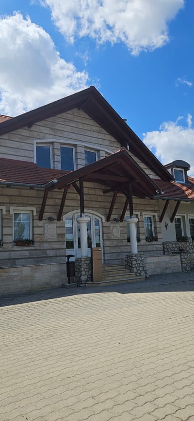 The image shows the facade of a building, possibly a public establishment or a large house, located in Cserszegtomaj, Hungary. The building is constructed with light-colored, horizontally laid stone blocks. It features a prominent, sloping, dark brown roof with red tiles. A covered entrance area is supported by thick wooden beams and pillars. The entrance itself consists of double glass doors. There are several windows on the upper levels, some of which are small and triangular. Decorative stone reliefs are visible on the building's exterior. The ground in front of the building is paved with light gray interlocking concrete blocks, forming a pathway or courtyard. To the left of the entrance, there is a dark gray trash receptacle. To the right of the entrance, a set of stone steps leads up to the building, flanked by stone walls and decorative plantings. The sky above is blue with scattered white clouds. The scene appears to be captured on a sunny day.