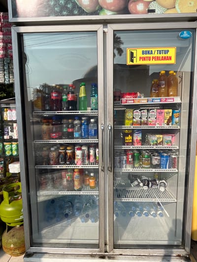 The image shows a double-door commercial refrigerator filled with various beverages. The refrigerator has a silver frame and glass doors. A yellow sign with red text reads "BUKA / TUTUP PINTU PERLAHAN," which translates to "OPEN / CLOSE DOOR SLOWLY." The top of the refrigerator has a picture of various fruits.

Inside the refrigerator, on the top shelves of the right side, there are several bottles of yellow liquid, likely juice. Below that, there are cartons of various milk drinks, including "ULTRA MILK" and "FRESH MILK." Further down are cans of drinks, some with colorful labels. The bottom shelves of the right side are filled with bottles of water.

The left side of the refrigerator contains bottles of soda, such as Fanta and Sprite, and other assorted drinks. There are also some packaged items on the lower shelves.

In the foreground, to the left of the refrigerator, there are two green propane gas tanks. One is larger and upright, while the other is smaller and on the ground.

The scene appears to be outdoors, possibly in front of a store. The lighting suggests it is daytime. The overall environment is that of a retail setting where drinks are sold.