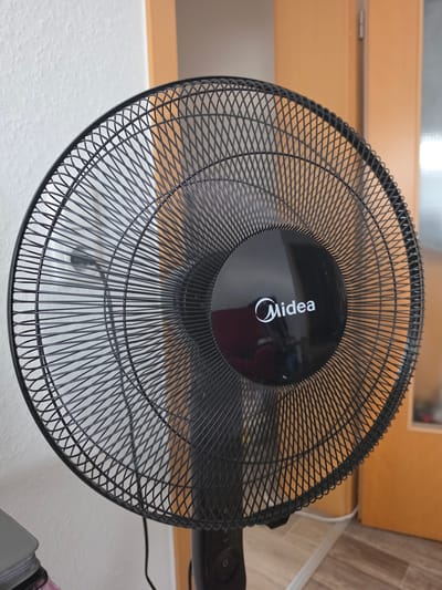 The image shows a close-up, overhead view of a black Midea brand electric fan. The fan's grille is intricate, with many thin wires creating a crisscross pattern. In the center of the grille, a circular black panel features the Midea logo, which consists of the word "Midea" in white text with a stylized white swirl above the "i". The fan is positioned in what appears to be an indoor setting, with blurred wooden furniture and a light-colored wall visible in the background. A white power cord is also visible extending from the right side of the fan, suggesting it is plugged in and ready for use. The lighting suggests it is daytime, but the indoor environment makes it difficult to ascertain specific weather conditions. No people are visible in the frame, and the activity implied is the fan's potential operation, though it appears to be stationary in the image. The setting offers no specific clues about Oberhausen, Germany, beyond being an indoor domestic or office space.