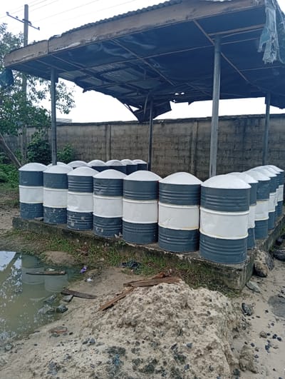 The scene in Igrita, Nigeria, presents a storage area containing approximately a dozen metal drums, painted with alternating gray and white bands, each capped with a white, dome-shaped lid. These drums are positioned on a raised concrete platform, sheltered by a corrugated metal canopy supported by metal poles. To the front of the platform is a pile of sand, with scattered debris around the area. There is a pool of water in the foreground. A concrete wall is partially visible behind the storage area. Vegetation is present on the left side of the frame. Power lines can be seen in the background above the structure.