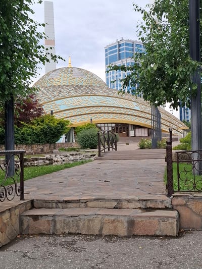 A paved path leads to the Argun Mosque, also known as the Mosque of the First President of the Chechen Republic Akhmad-Khadzhi Kadyrov, in Argun, Russia. The mosque features a large, ornate dome with Arabic inscriptions and a golden crescent moon on top. A tall, white minaret stands to the left of the dome. The path is made of stone slabs and is lined with decorative metal railings and lush green bushes. In the background, a modern blue glass skyscraper rises above the trees. The sky is overcast, suggesting a cloudy day.