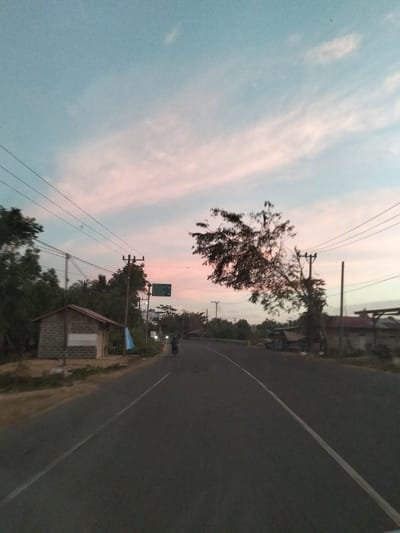 The image depicts a two-lane asphalt road at twilight in Pantonmakmur, Indonesia, with a light blue sky transitioning to pink at the horizon. The road extends into the distance, curving slightly to the right, marked by a solid white line on the right and a broken white line on the left. On the left side of the road, in the foreground, stands a small, single-story building constructed of light-colored blocks with a dark, sloped roof. Utility poles with multiple power lines run parallel to the road on both sides. A green rectangular road sign is visible on a utility pole on the left, and a light blue banner or cloth hangs nearby. Further down the left side, dense green foliage is present. On the right side of the road, trees with dark foliage are visible, with one tree featuring sparse branches extending over the road. Indistinct structures are visible through the trees on the right. A single individual on what appears to be a motorcycle or bicycle is observed traveling away from the viewer in the left lane of the road. The overall lighting is dim, consistent with dusk or dawn.