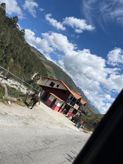 The image depicts a steep, mountainous landscape in Mucurubá, Venezuela, with a road in the foreground. A two-story building with a red tiled roof and white and red walls is situated on the right side of the road. A fence runs along the left side of the road, bordering a steep, vegetated hillside. A horse with a saddle is standing on the side of the road near the fence, facing left. The sky above is blue with scattered white clouds. The image is taken from a slightly tilted perspective, suggesting it was captured from a moving vehicle.