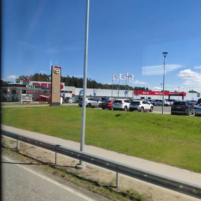 The image captures a commercial area on a sunny day, likely in Berģi, Latvia, with a clear blue sky and scattered clouds. In the foreground, a grey metal guardrail separates a paved road from a grassy area. The grassy area is dotted with yellow wildflowers. Behind the grass, a white paved walkway runs parallel to the road.
The main focus of the scene is a commercial complex featuring a gas station and a car dealership. The gas station, labeled "KOOL," has multiple pumps under a green and white canopy. Next to it, a tall, wooden sign with a red top displays the Toyota logo and the words "Toyota Servis," "Approved," and "Toyota Professionals" in white text. Further to the right, a white building with red trim features large text reading "Būvel" and "Dārzam," suggesting it may be a construction or garden supply store. Additional white flags with blue emblems are visible, some bearing the word "WESS."
Several cars are parked in front of the commercial buildings, including a silver SUV, an orange sedan, a white SUV, and a dark-colored SUV. A few people are partially visible, including one standing near a car on the far right and another person in a blue checkered shirt on the left side of the white building. The background is filled with a dense line of trees. The overall impression is of a modern commercial hub situated in a suburban or semi-rural environment.