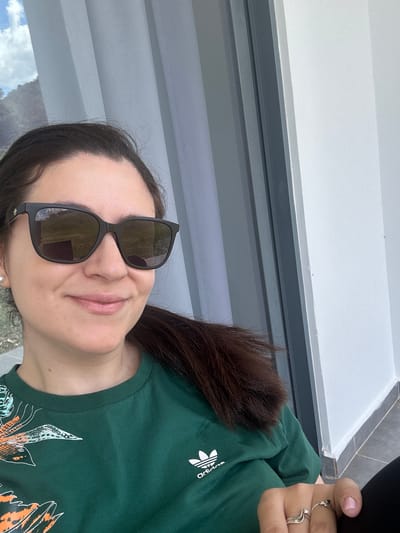 A woman wearing dark sunglasses and a dark green Adidas shirt with a floral pattern is holding a phone in her right hand. She is standing in front of a window with a white sill and a grey frame. The background seen through the window suggests an outdoor area with vegetation and possibly a distant view of Faliraki, Greece. The image is rotated counter-clockwise.