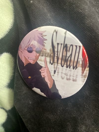 A round, glossy button featuring a cartoon image of a man with white hair and sunglasses, pointing with his left hand. The background of the button includes the word "Shibuya" written vertically in a stylized font, and a blurred depiction of an urban area. The button rests on a dark, textured fabric. No location in Whitby, Canada, is depicted in this image.