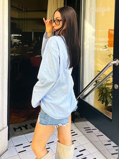 The image shows a young woman standing in front of a doorway. She has long, dark hair, is wearing glasses, and is dressed in a light blue oversized sweatshirt and denim shorts. She is also wearing tan boots. The doorway is glass and has a metal handle. The woman is looking to the side and adjusting her glasses. The background shows what appears to be a restaurant or shop interior. There is a mat on the floor inside the doorway with the letters "MY.B" visible. The floor outside the doorway is tiled. The scene does not contain any elements that would suggest a specific location within the city of Korostyshiv, Ukraine.