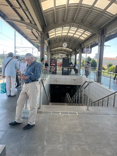 A group of people are standing on a train platform in Zeytinburnu, Turkey, under a covered walkway. In the foreground, a man in a blue plaid shirt and beige pants is looking at his phone. Behind him, other people are waiting, some with bags. A set of stairs leads down from the platform. The environment appears to be a modern transit station with metal and glass elements. The sky is visible in the background, indicating it is daytime.