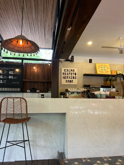 The image shows the interior of a cafe, likely located in a small town in Costa Rica. The cafe is decorated with a wooden ceiling and a white tiled counter. A wicker chair sits on the tile floor with a menu board behind it. The cafe has a large sign on the wall reading "Calma, Respeto, Abrazos, Amor," likely a motto or slogan. There are two other signs behind the counter, which appear to be advertising food or drinks. The cafe has an espresso machine on the counter, indicating the preparation of coffee drinks. The location coordinates correspond to an area in Santa Fe, a town in the province of Alajuela, Costa Rica.