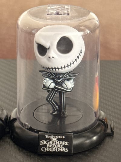This media file shows a close-up, vertically oriented shot of a Jack Skellington figurine from "The Nightmare Before Christmas." The figurine is encased in a clear plastic cylindrical container with a black base and lid. Jack is depicted in his iconic black and white pinstripe suit, with his skull-like head prominent. His arms are crossed in front of him.
The background consists of a textured, dark surface, possibly a desk or table. The lighting is direct, creating some highlights on the plastic container and the figurine. The overall mood is that of a collectible item displayed.
Visible text on the black base reads "TIM BURTON'S THE NIGHTMARE BEFORE CHRISTMAS" in a stylized font. The context provided indicates the location is Vespolate, Italy, though this is not directly evident from the image itself. No specific time of day or weather conditions can be inferred. The figurine itself is the sole subject, and no particular activity or event is taking place beyond its static display. Notable details include the stitched detailing on Jack's face and the distinct pattern of his suit.