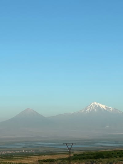 This is a landscape photograph taken in Zangakatun, Armenia, depicting the prominent twin peaks of Mount Ararat under a clear blue sky. The left peak is a conical, snow-capped mountain, while the right peak is taller and also snow-capped, with more rugged terrain visible. In the middle ground, there are bodies of water, possibly a lake or reservoir, and a small town or village with buildings. In the foreground, there is dry vegetation, fields, and a solitary electrical pylon. The image captures a serene, natural environment with human infrastructure present.