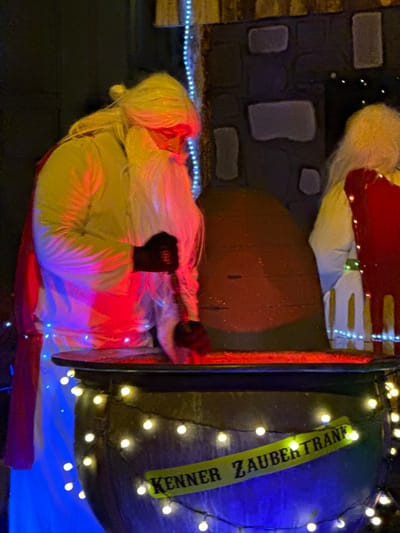 The scene depicts a festive display in Wasserbillig, Luxembourg, observed at night or in low light conditions. The primary subject is a costumed figure on the left, resembling a Santa Claus or wizard, with a long white beard, a white robe, and a red cape. This individual wears black gloves and is actively stirring a large, dark cauldron in the foreground with a long implement. The cauldron's contents emit a red-orange glow, and the figure's face is illuminated by a red light. The lower portion of the figure and the cauldron are lit with blue light, while warm white string lights are draped around the cauldron. A yellow sign partially visible on the cauldron reads "KENNER ZAUBERTRANK." To the right, a second costumed figure, similarly adorned with white hair and a white robe with red and gold accents, is partially visible. The background features a dark, textured wall, possibly made of stone or a stone-like material, and a large, dark, rounded structure behind the cauldron. Blue string lights are visible vertically along the left side of the background wall.