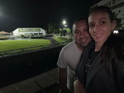 Captured at night in the Borough of Spelthorne, United Kingdom, this image shows a couple posing for a selfie. The woman, on the right, is smiling. To her left, a man is also smiling. They are positioned in front of a tiered landscape featuring a grassy area enclosed by a low wall. In the background, a white building with a sign that reads "PROMISE THE EARTH WILL SHAKE" is visible. A row of trees and streetlights stretches along the backdrop. The area seems well-lit.