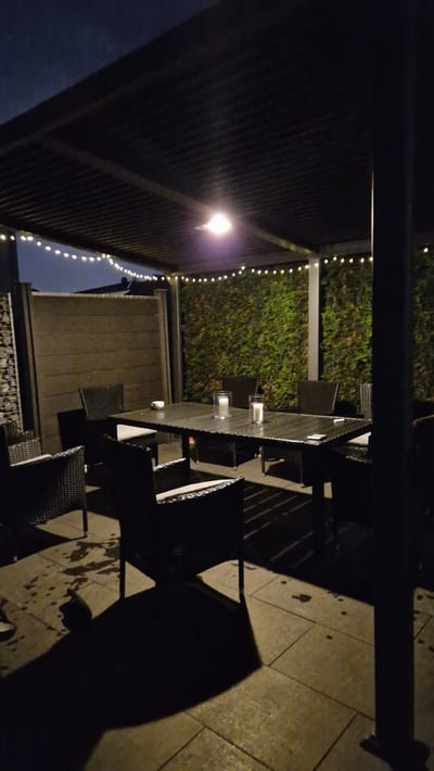The image depicts an outdoor patio area at night, likely a backyard or restaurant seating area, located in Ichenhausen, Germany. A dark, slatted roof structure is supported by a thick pillar on the right side of the frame, with string lights lining the underside of the roof. A central dining table, made of a dark material, is set with two tall glass cylinders containing candles, one of which is lit. Several chairs with dark woven wicker frames and light-colored cushions surround the table. The patio floor is paved with large, square tiles, which appear wet, suggesting recent rain. To the left, a dark wooden fence extends horizontally, and to the far left, a section of a stone wall is visible. Behind the table, a dense green hedge forms a backdrop. The overall lighting is dim, with the primary illumination coming from a light fixture on the roof and the candle. No people are present in the scene.