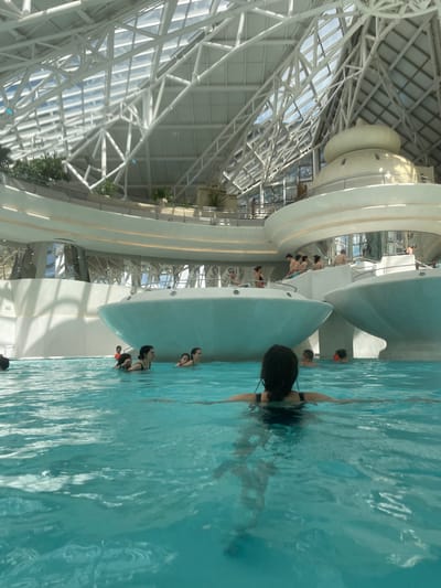 The image captures a scene within the Caldea Thermoludic Spa in Les Escaldes, Andorra. The spa features a distinctive architectural design with a large, white, metal-framed glass dome. Inside, a series of interconnected pools and water features are visible. Multiple people are present in the pools, some swimming, others standing and interacting near the edges. The water appears clear and blue, creating a vibrant contrast with the white structures of the spa. The photo shows a swimming pool with people in it, inside of a building that has a white dome ceiling.