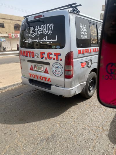 This image captures a daylight street scene in Rijiyar Lemo, Kano, Nigeria. A silver Toyota Hiace minivan, license plate KANO 9SR-400ZL, is the primary subject, viewed from its rear-passenger side while stationary on a paved asphalt road.
The van features a roof rack and numerous decals. The rear window is largely covered by a black sticker with white Arabic calligraphy. Below this, the red text "NARTO F.C.T." is present, along with a circular logo for the "NATIONAL ASSOCIATION OF ROAD TRANSPORT OWNERS (NARTO) FCT BRANCH" on the right rear panel. The word "TOYOTA" in red is affixed to the lower rear bumper, which displays scuffing and minor impact damage on the passenger side. The rear passenger side window also has a black sticker with gold Arabic calligraphy, and below it, the red text "MASHA ALLAH" and "SPORT" are visible on the van's body. The rear passenger side wheel is a black steel rim.
The asphalt road beneath the van shows cracks and scattered light-colored debris. A distinct shadow, cast by the van and suggesting strong overhead sunlight, extends from the vehicle onto the road. A concrete curb separates the road from a paved sidewalk. In the background, across the sidewalk, are light-colored, multi-story buildings, one of which displays a sign reading "YUSR SUPERSTORES". A single, indistinct individual in dark clothing is partially visible on the distant sidewalk. In the extreme right foreground, a pink object with a "GUCCI" logo and a partial reflection of a human face are visible, indicative of a reflective surface belonging to the observer's immediate vicinity.