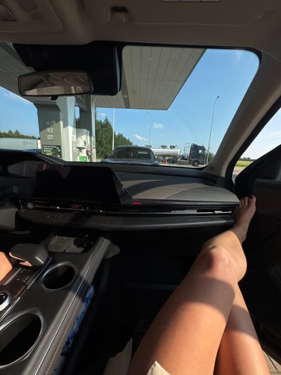 The image, taken from inside a car at a gas station in Novoe Mart'yanovo, Russia, shows a person's legs and feet propped up on the dashboard. Outside the car, a gas pump with prices indicated as 95 and 92 is visible, along with a fuel truck and a clear blue sky. The car's interior features a modern dashboard with a large screen and a console with cup holders. The sunlight casts a warm glow on the scene.