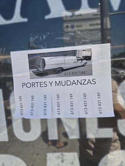 A torn flyer is affixed to a glass surface, possibly a shop window or a bus shelter, in Collado Villalba, Spain. The flyer features a black and white image of a van and the text "PORTES Y MUDANZAS" (Spanish for "Moving and Removals"). Below this text, a perforated strip contains multiple copies of a phone number, "613 431 149". The flyer is partially obscured by tape and reflections on the glass. The background shows a blurred urban environment with shadows and faint lettering on a dark surface.