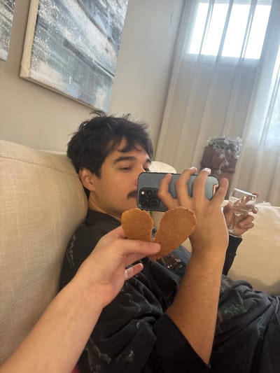 An indoor scene in the city of Menashe Regional Council, Israel, features a male individual with dark hair and a mustache, reclining on a light beige textured couch. He is wearing a dark, possibly patterned, shirt. His right hand holds a dark-colored smartphone with a triple-lens camera array, aimed towards the foreground. His left hand holds a clear stemmed glass containing liquid. In the foreground, another person's arm and hand are visible, holding two light brown, flat, irregular cookie-like objects, positioned in front of the smartphone. The background shows a light-colored wall with two abstract paintings in shades of white, grey, and blue. A large window with white vertical bars and sheer curtains is visible, along with a dark, ornate pot containing a white decorative plant or item.