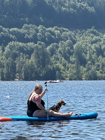A woman and a dog are on a paddleboard in Lake Titisee, Titisee-Neustadt, Germany. The woman is wearing a black swimsuit and has a pink bandage on her upper arm. The dog is wearing a harness. In the background, there is a wooded hillside. The water is blue and choppy with sunlight reflecting off it. Several other boats and paddleboards are visible in the distance.