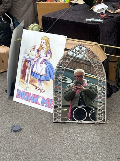 The image depicts a collection of objects strewn on a pavement or street in St Albans, United Kingdom. A mirror, reflecting a person taking a selfie, lies at an angle. The mirror has a silver, ornate frame. A white sign with an illustration of Alice from Alice in Wonderland and the text "DRINK ME" is also present. The scene appears to be part of an outdoor market or sale, with other items like boxes, a black draped table, and other unidentified objects scattered around. The ground is asphalt. A discarded can lid is visible on the asphalt.